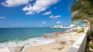 Uma praia com águas turquesa cristalinas, um litoral rochoso e um edifício branco ao fundo.