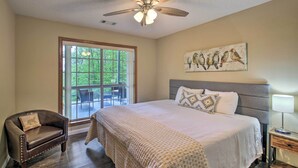 Une chambre avec un grand lit, un ventilateur de plafond, une fenêtre donnant sur un paysage verdoyant, un fauteuil et un tableau représentant des oiseaux.