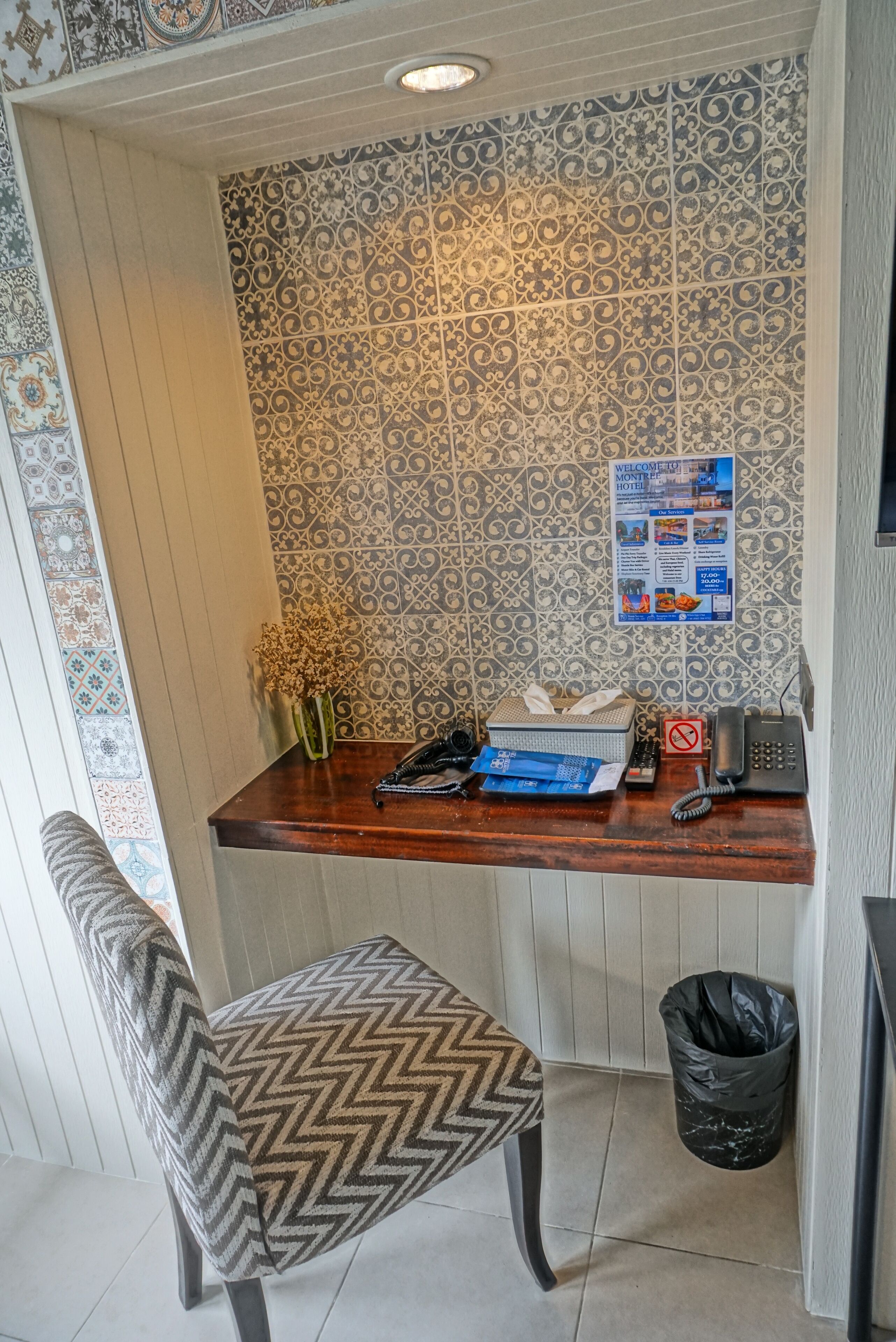 Un comptoir d’accueil avec un mur orné d’un motif, un vase contenant des fleurs séchées, un téléphone, une poubelle et une chaise à motifs chevrons.