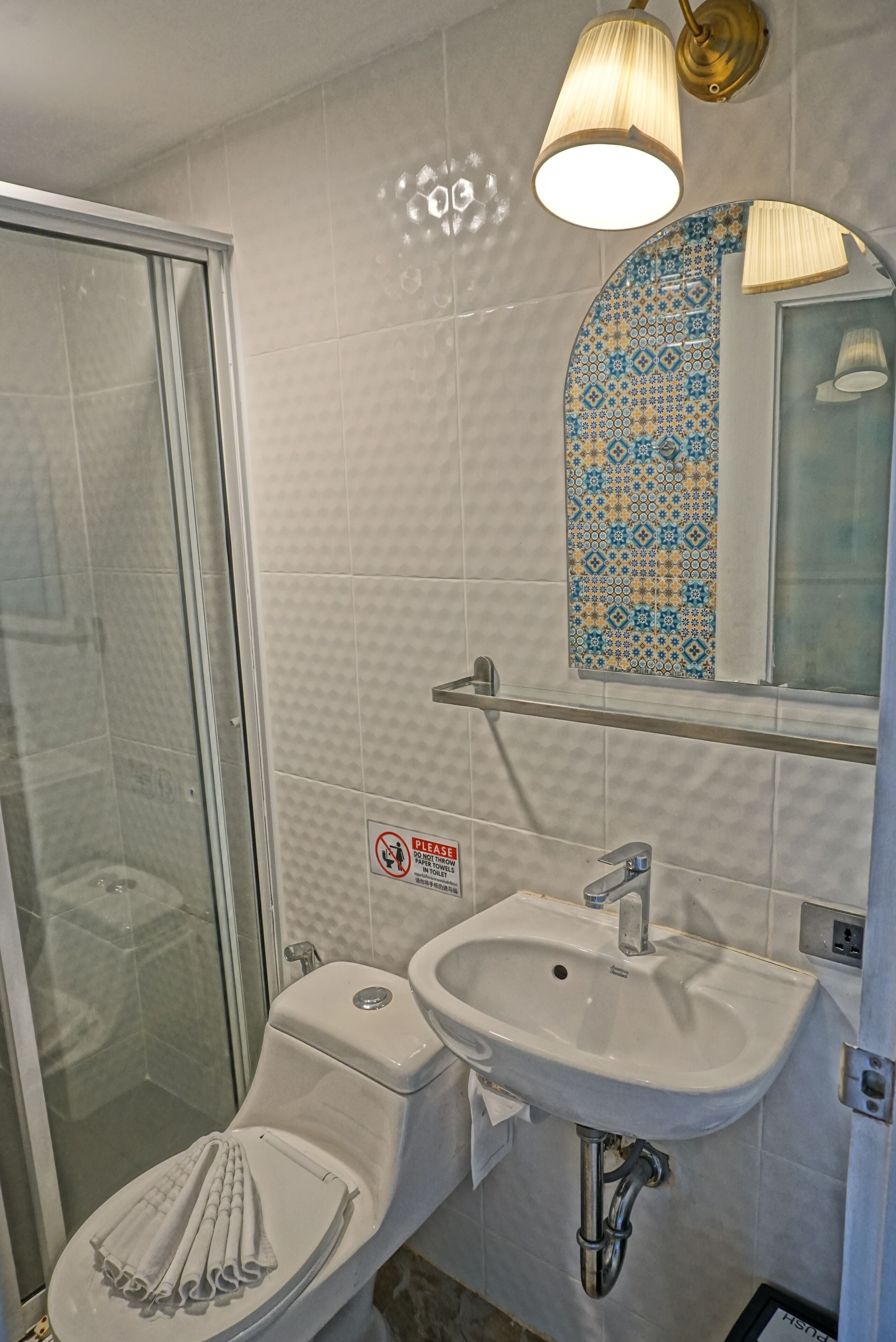 Baño con cabina de ducha de vidrio, inodoro con toalla, lavamanos y espejo con azulejos estampados.