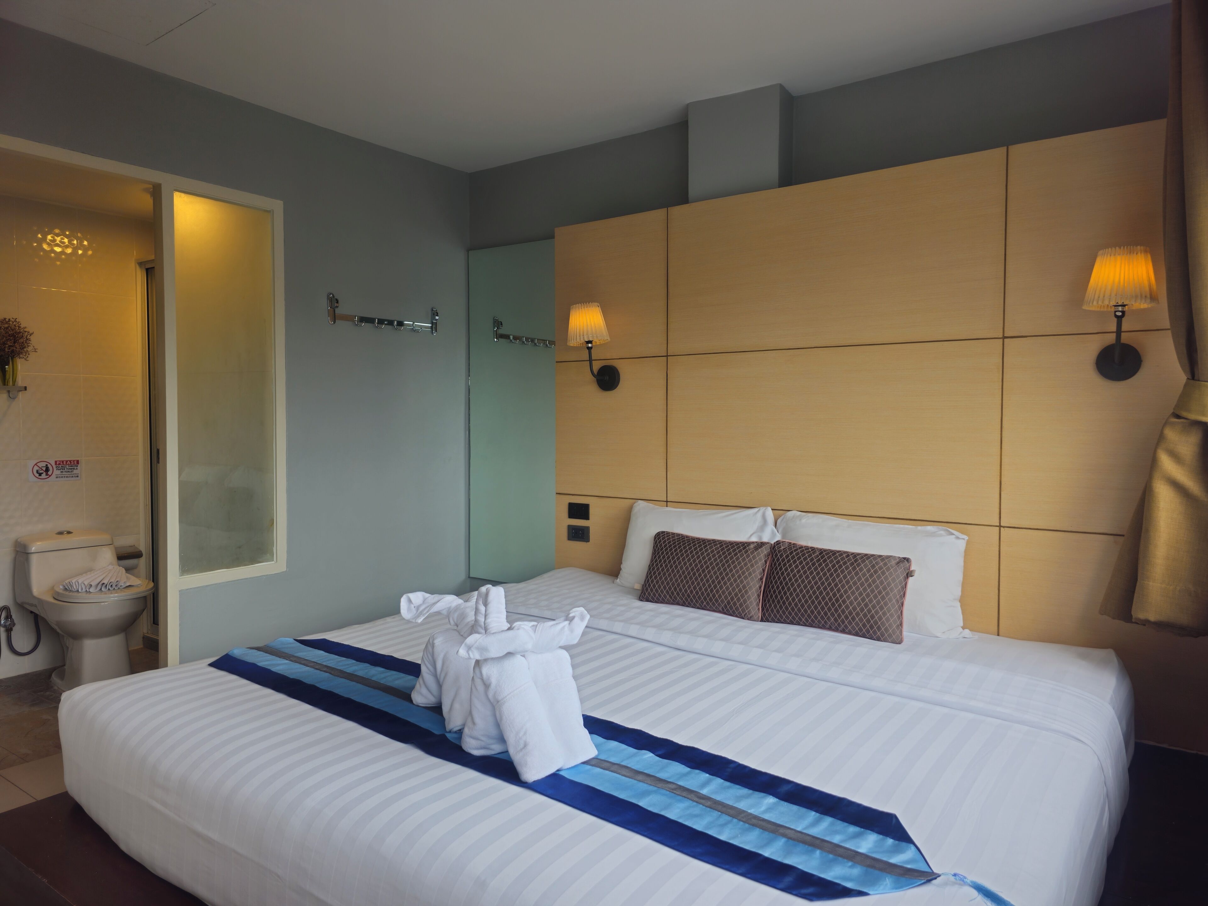 A hotel room with a large bed, a bedside table, a nightstand, a wall-mounted lamp, and a bathroom with a toilet and sink.