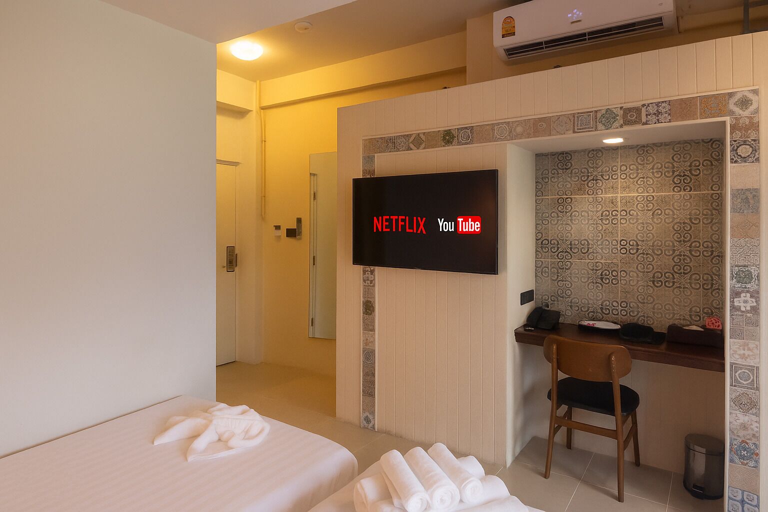 A hotel room with a bed, a desk with a chair, a TV displaying Netflix and YouTube logos, and a bathroom door.