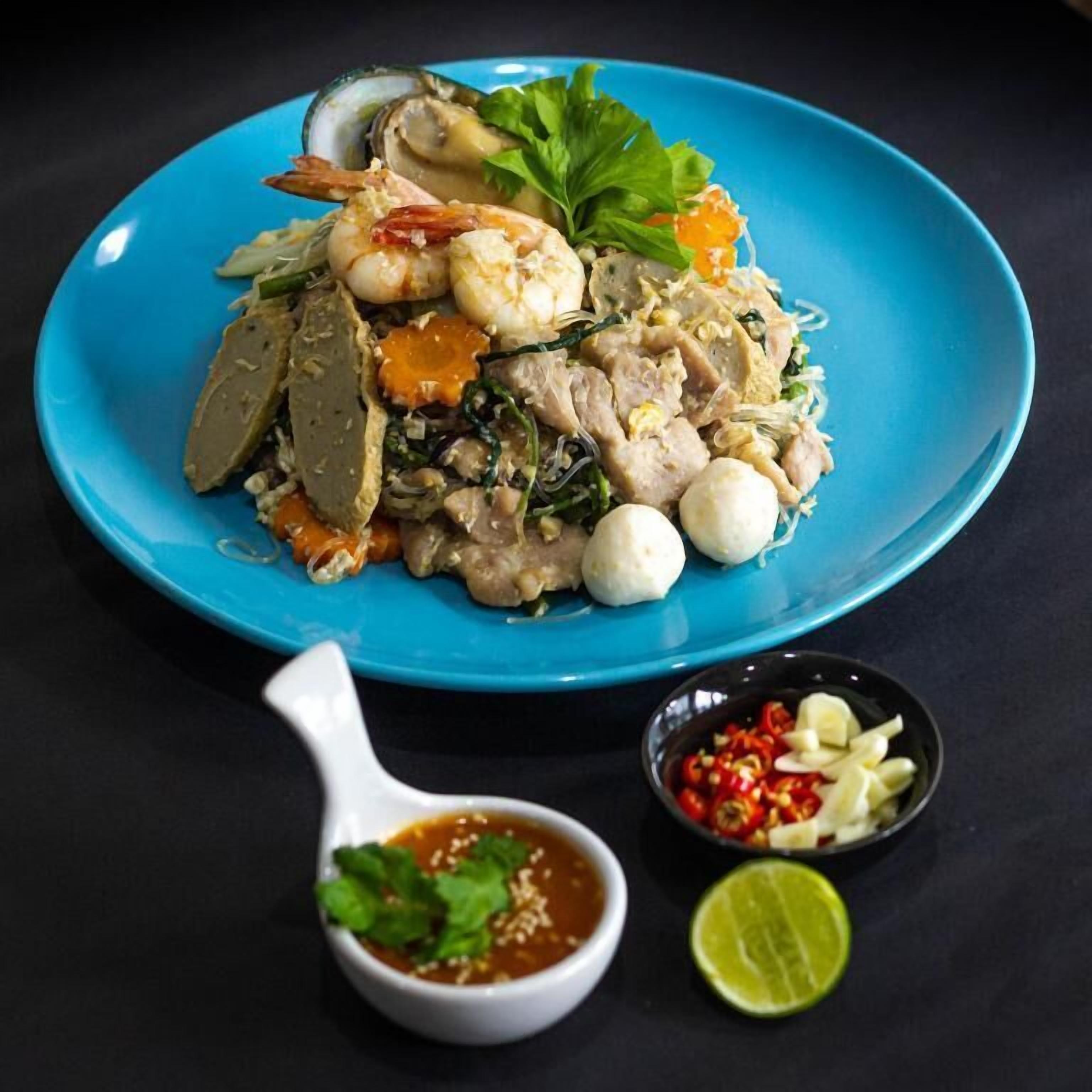 Un plato de pad thai con camarones, fideos y verduras, acompañado de un tazón pequeño de salsa y chiles en rodajas.