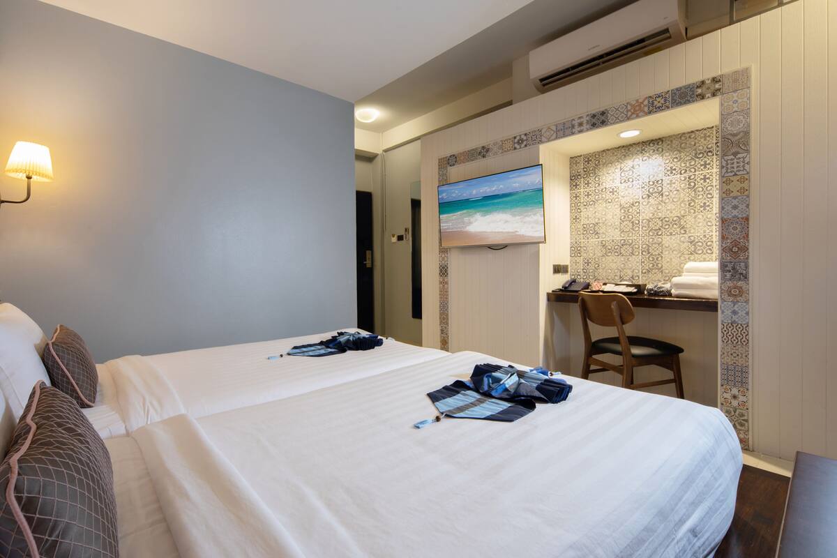 A hotel room with two beds, a desk, a chair, a TV, and a patterned wall.