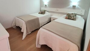 Habitación doble con dos camas, cada una con una colcha blanca y un cojín estampado. Hay una mesita de noche entre las camas y el piso es de madera.