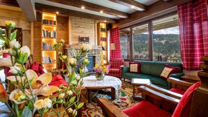 A cozy living room with a fireplace, bookshelves, a green sofa, and a large window offering a scenic view. - La Noi Guesthouse (Grevena)