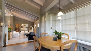 A dining area with a wooden table and chairs, a chandelier, and a view through large windows with blinds. - Serene Seaside Bliss - Lush & Secluded Garden Oasis (Kalamata)