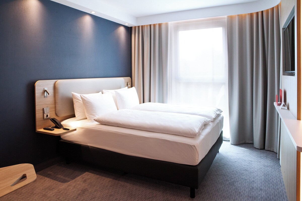 A modern hotel room with a large bed, a bedside table with a phone, and a window with curtains.