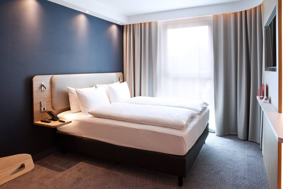 A modern hotel room with a large bed, a bedside table with a phone, and a window with curtains.