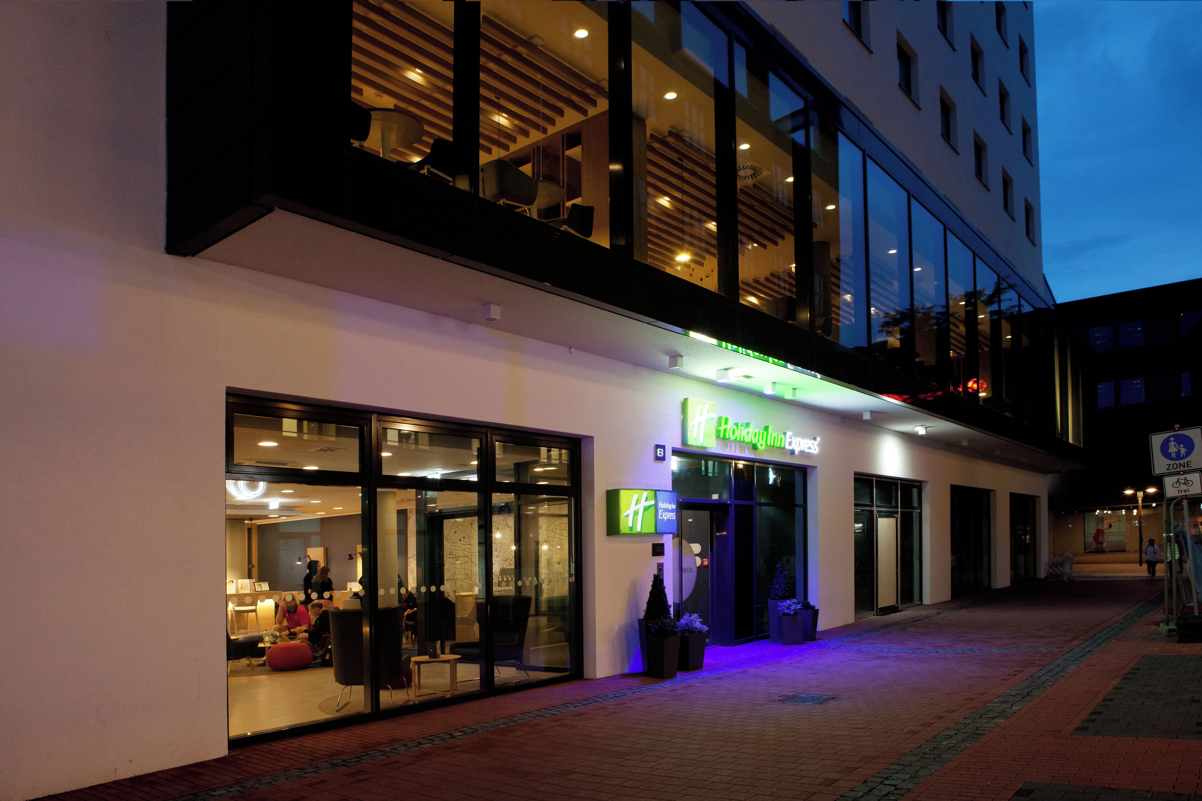 A Holiday Inn Express hotel with a glass facade, outdoor seating area, and a well-lit entrance.