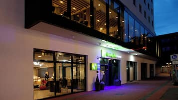 Un hotel Holiday Inn Express con fachada de vidrio, zona de estar al aire libre y una entrada bien iluminada.