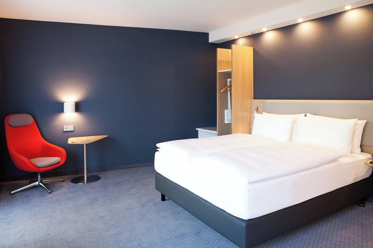A modern hotel room with a large bed, a red chair, a small table, and a wall-mounted light.