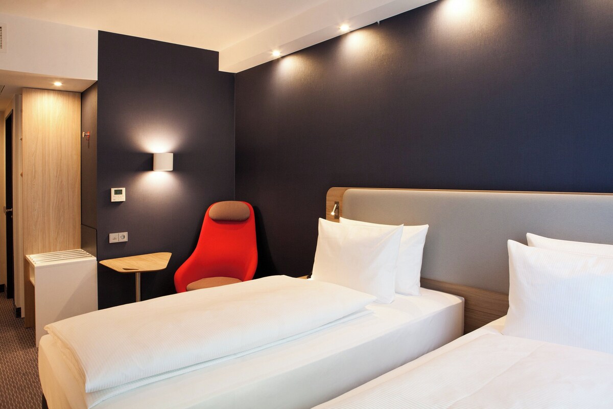 A hotel room with two beds, a red chair, a small table, and a wall-mounted light.