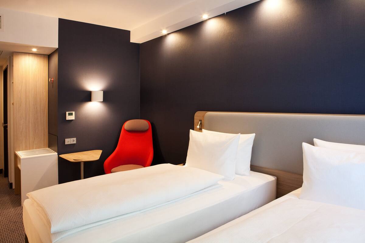 A hotel room with two beds, a red chair, a small table, and a wall-mounted light.