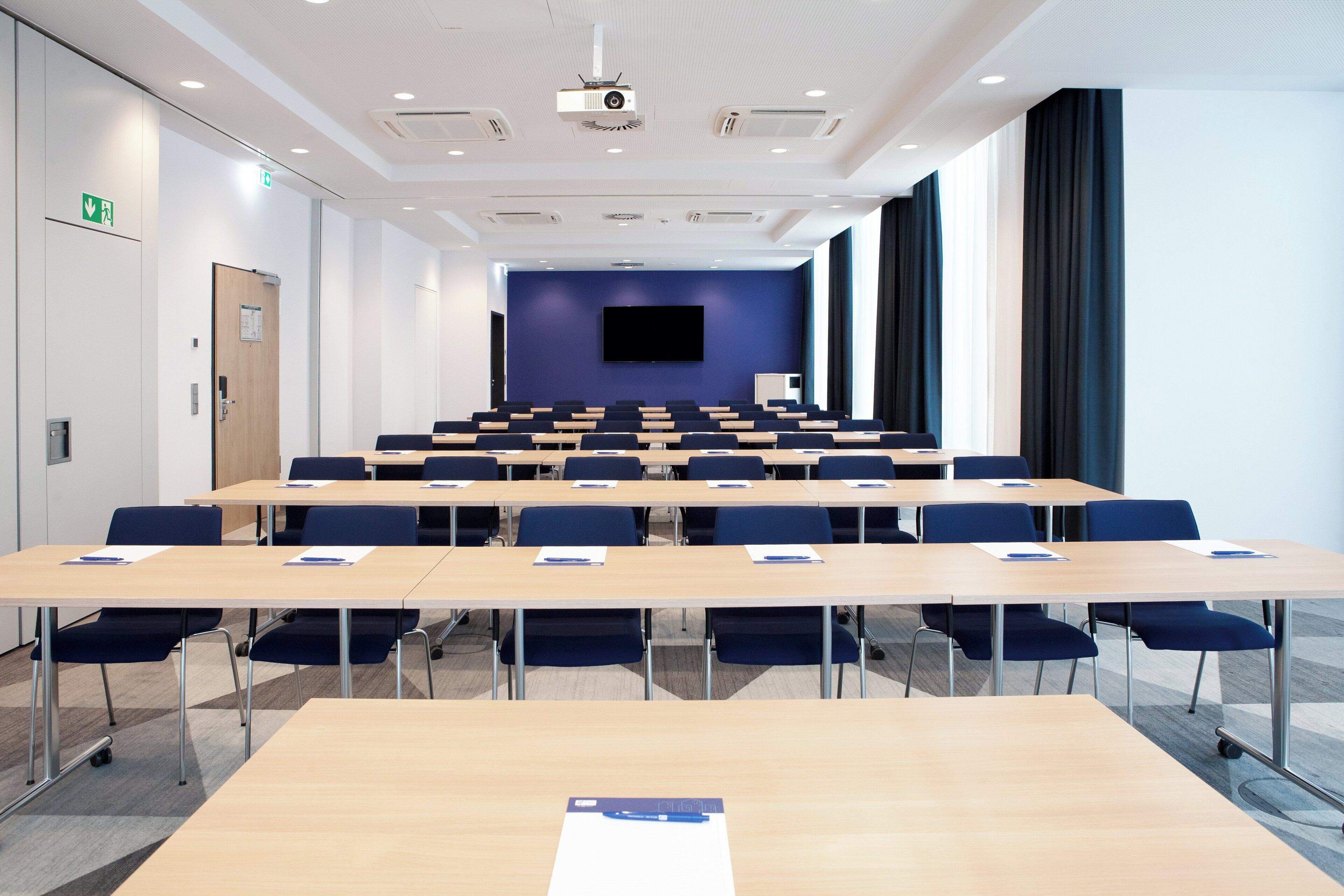Une salle de conférence avec des rangées de tables et de chaises, un projecteur et un grand écran.