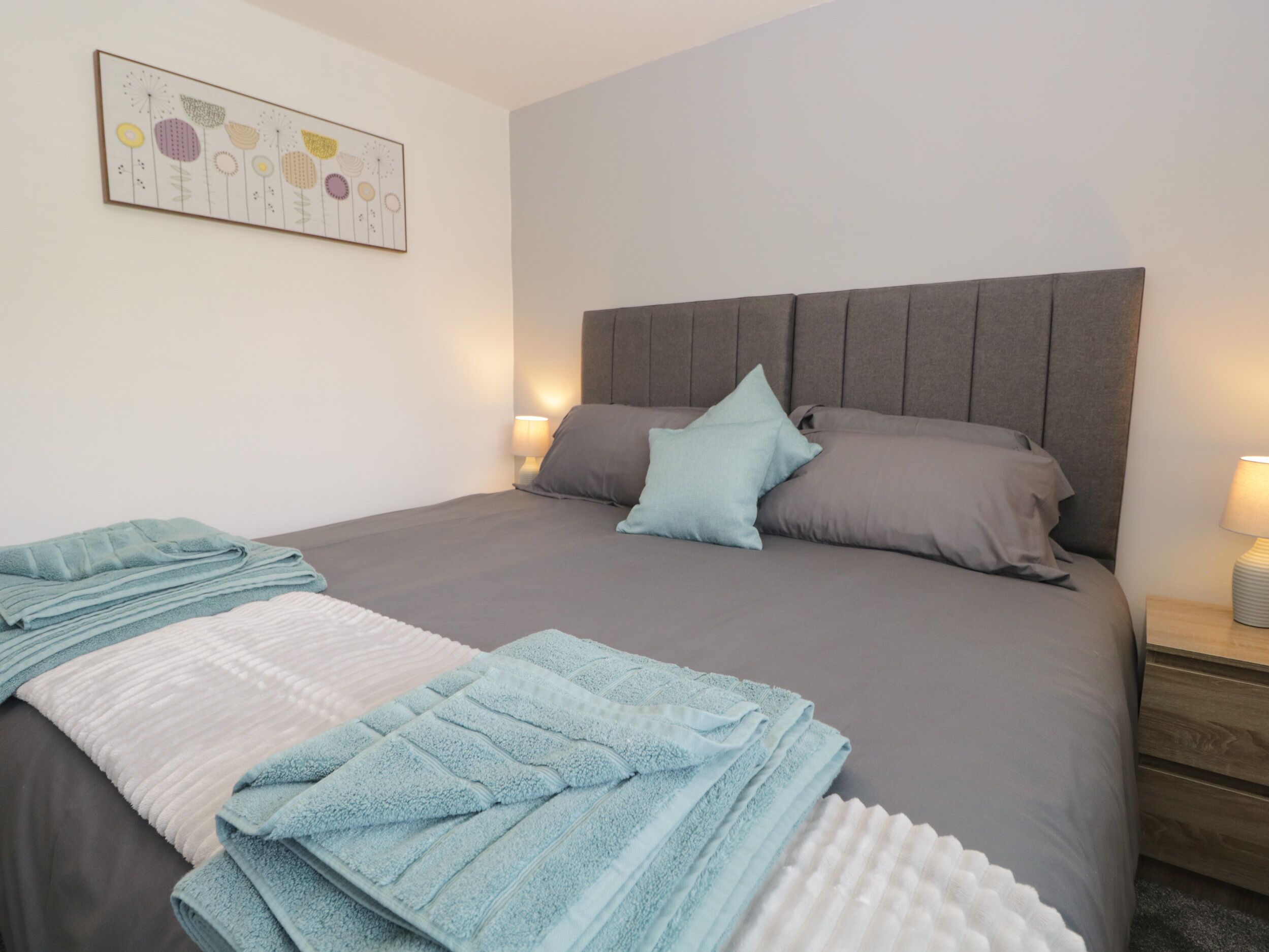 Una cama bien hecha con ropa de cama gris y azul, dos lámparas de noche y una obra de arte en la pared sobre la cama.