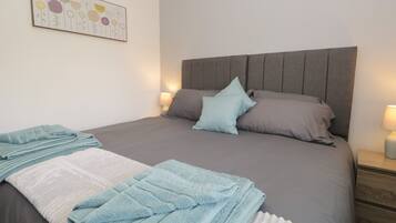 Una cama bien hecha con ropa de cama gris y azul, dos lámparas de noche y una obra de arte en la pared sobre la cama.