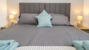 A neatly made bed with a grey headboard, two bedside lamps, and a folded towel on the floor. - 26 Hycemoor Way (Millom)