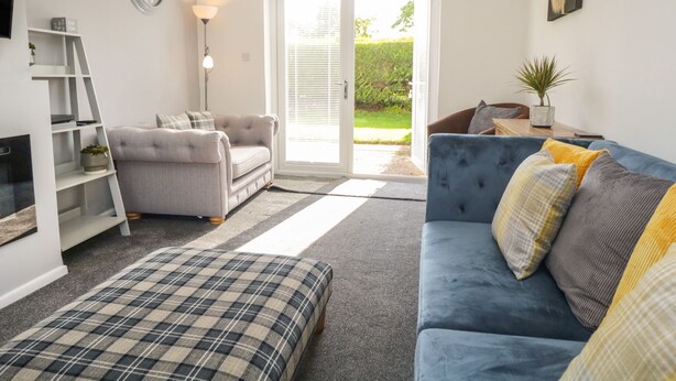 A living room with a grey sofa, a blue sofa, a plaid ottoman, a white shelving unit, and a door leading to a garden. - 26 Hycemoor Way (Millom)