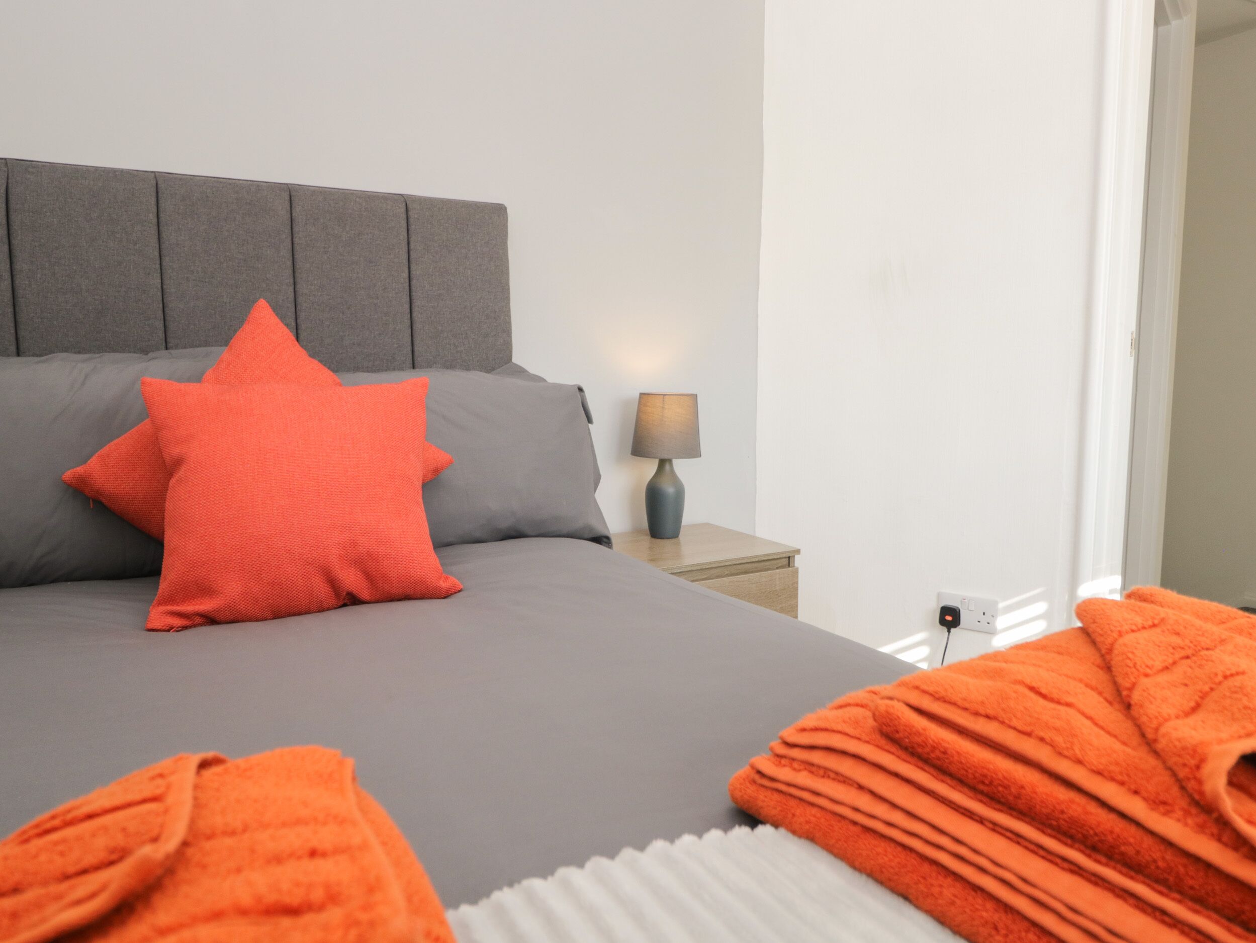 A neatly made bed with grey bedding and a red pillow, a bedside lamp, and a folded orange towel on the nightstand.