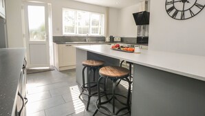 A modern kitchen with a large island, two bar stools, a clock, and a window with blinds. - 26 Hycemoor Way (Millom)