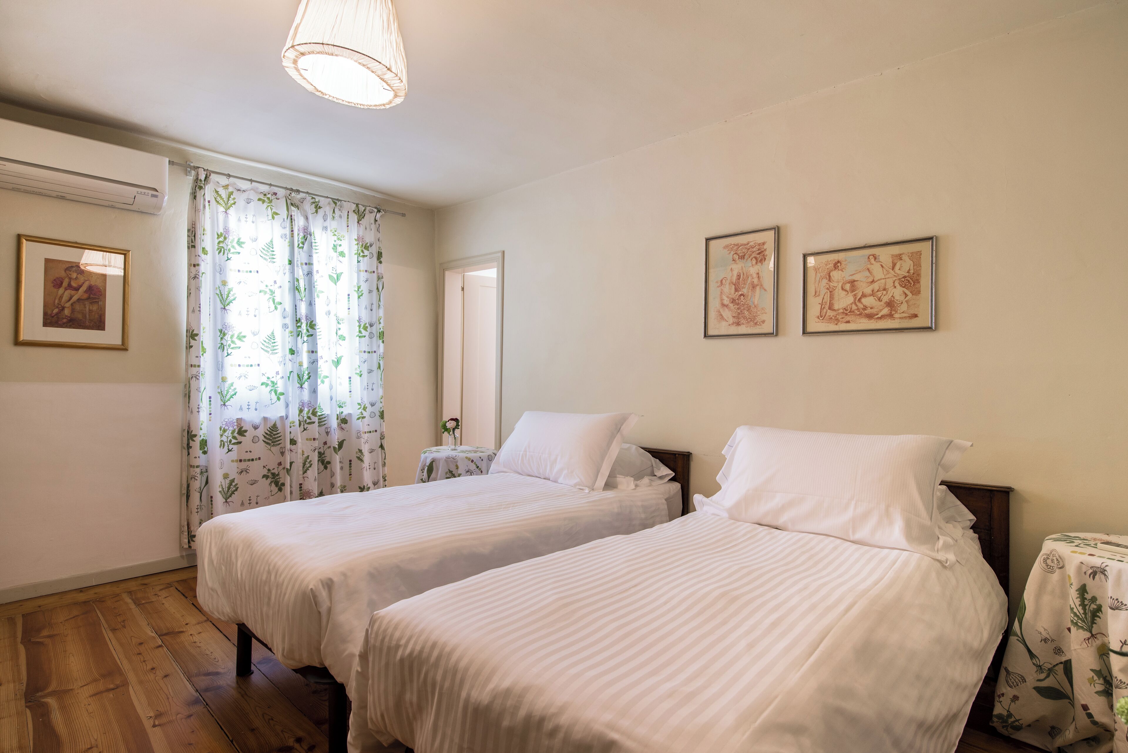 A bedroom with two beds, a window with curtains, a bedside table, and wall art.