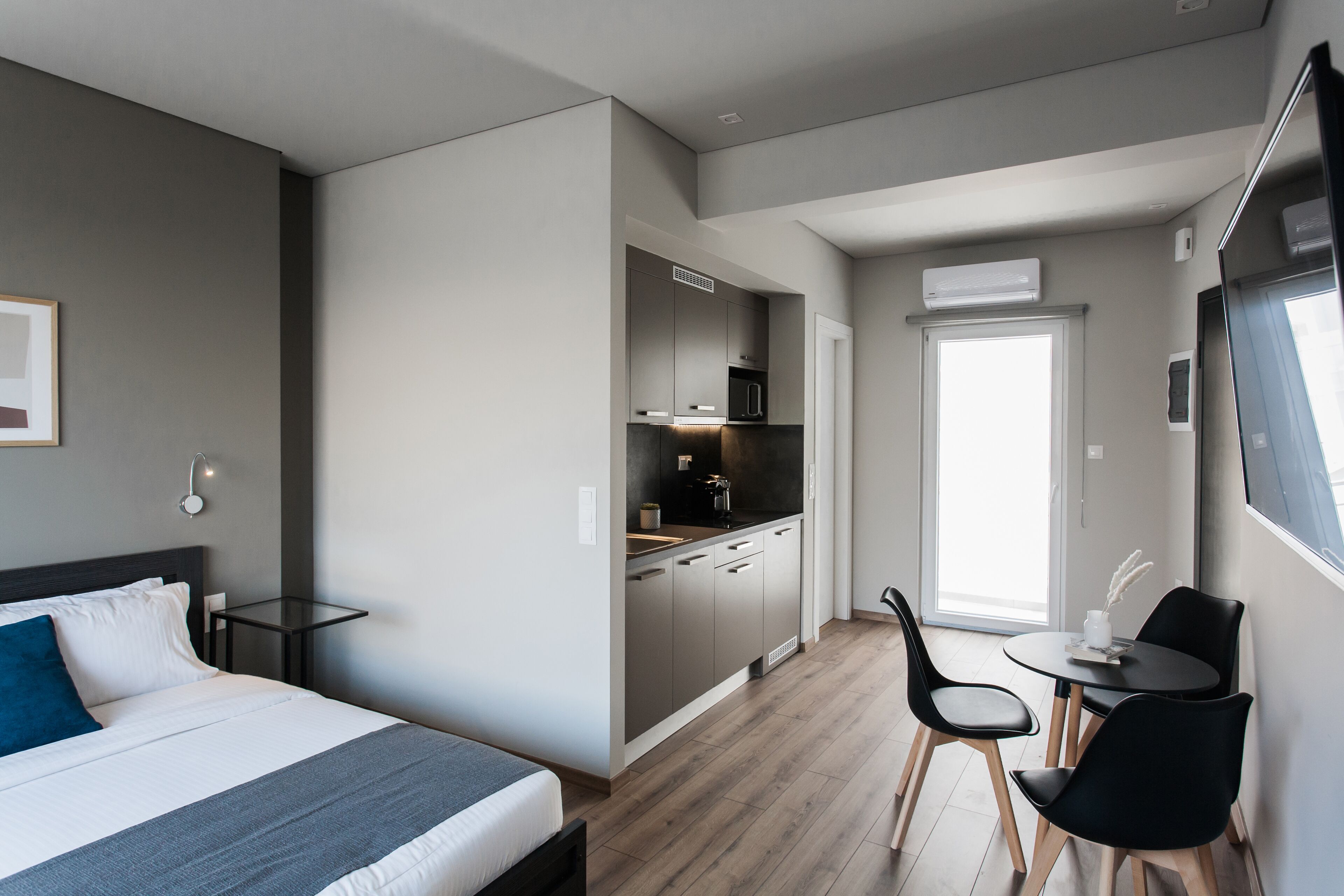 A modern hotel room with a bed, a small kitchenette, a dining area with chairs, and a large window.