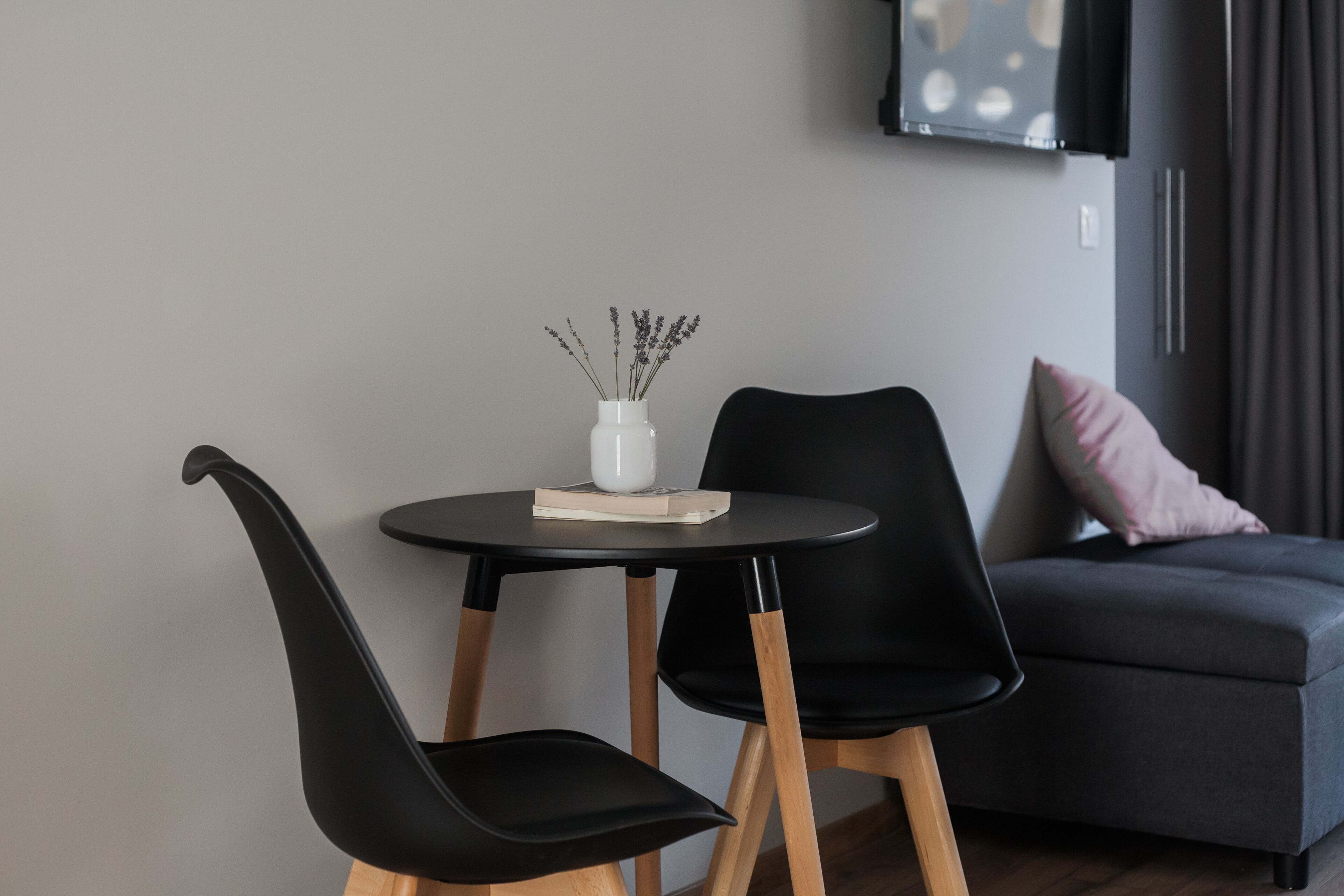 Una sala de estar moderna con una mesa redonda, dos sillas negras, un jarrón blanco con flores secas, un libro, un sofá gris con un cojín rosa y un televisor de pantalla plana montado en la pared.