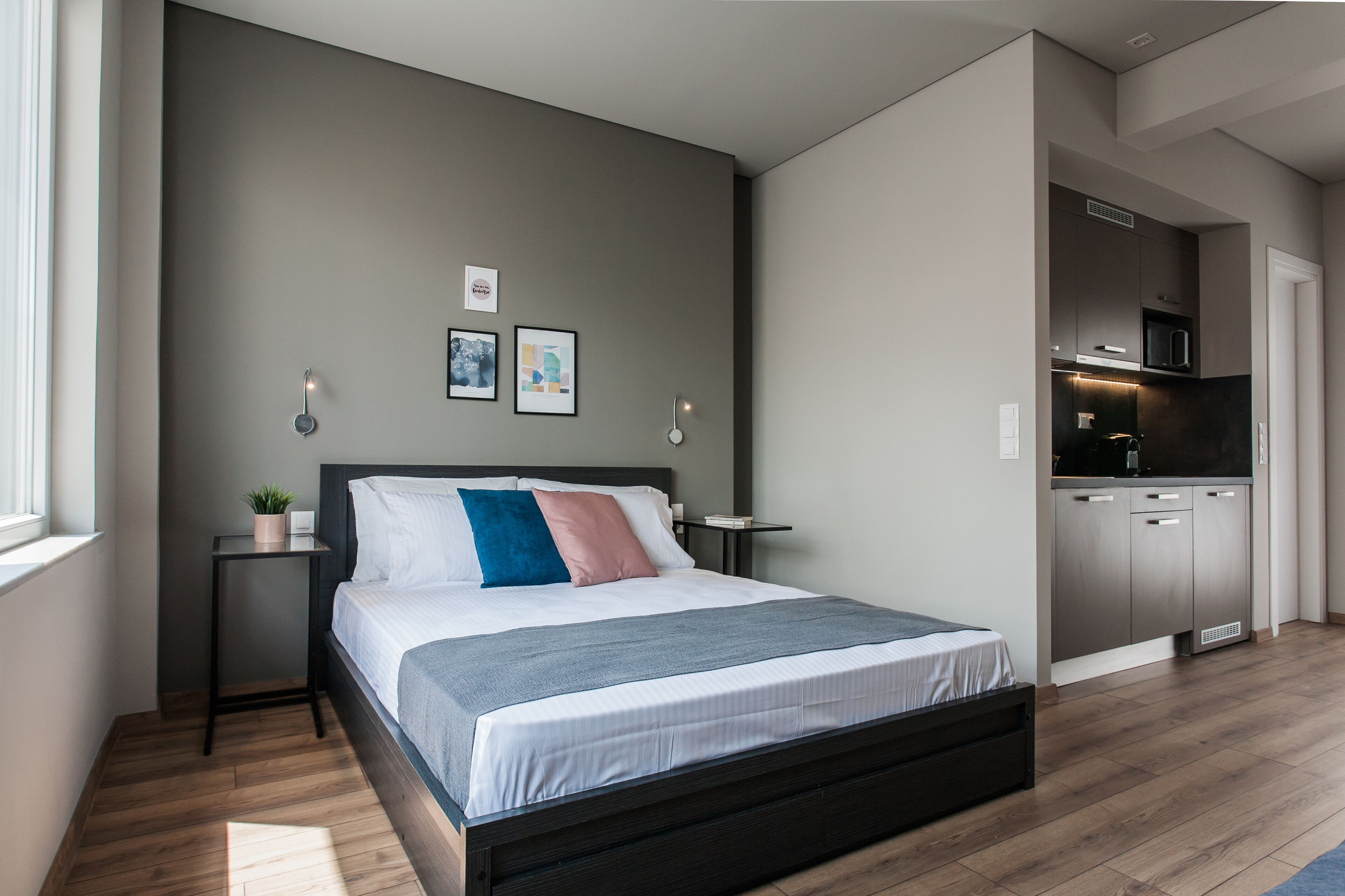A modern bedroom with a large bed, bedside tables, a small plant, and a view into the adjacent kitchen.