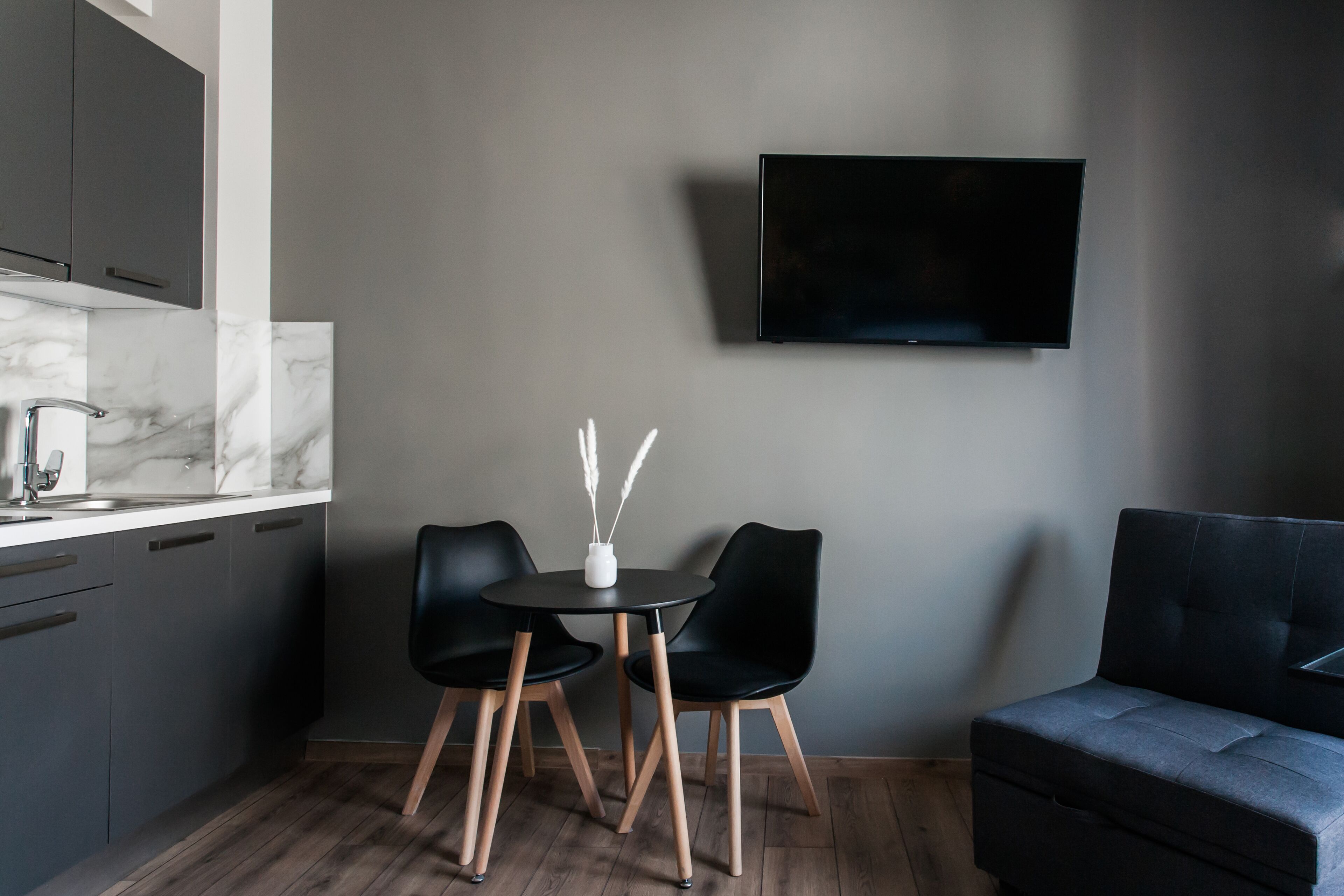 Une cuisine moderne avec des meubles de couleur sombre, un plan de travail en marbre et une table à manger noire avec deux chaises. Un téléviseur à écran plat est fixé au mur au-dessus de la table.