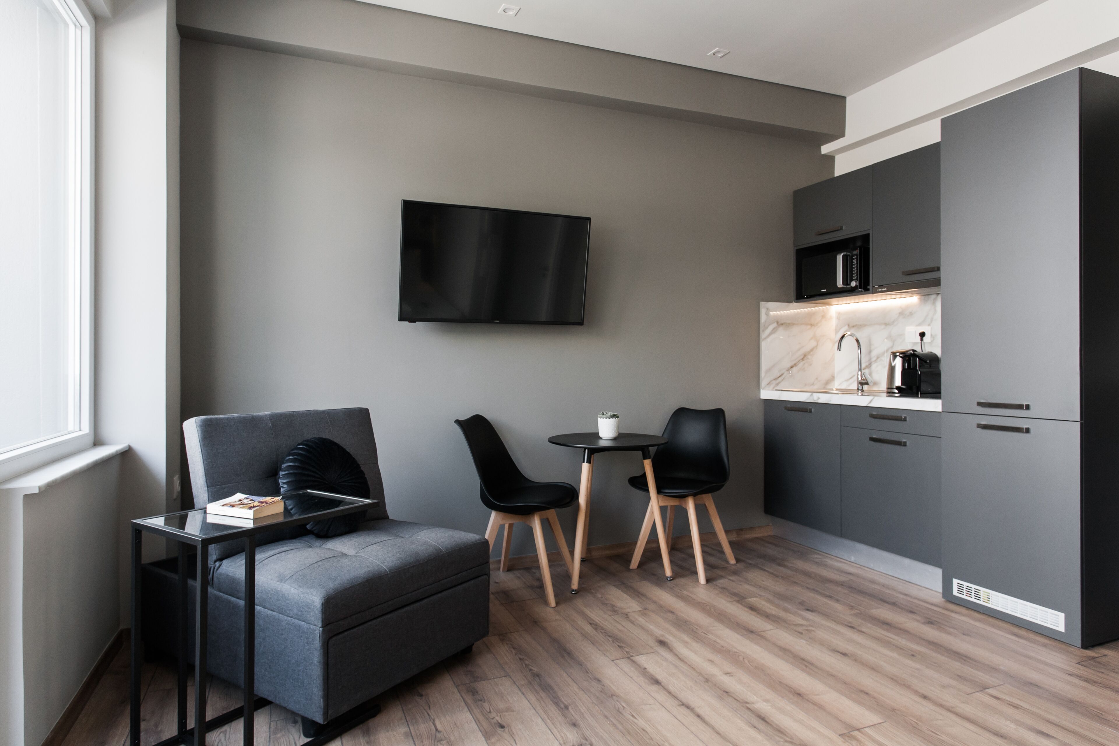 A modern kitchen with grey cabinets, a flat-screen TV, and a dining area with two black chairs and a small round table.