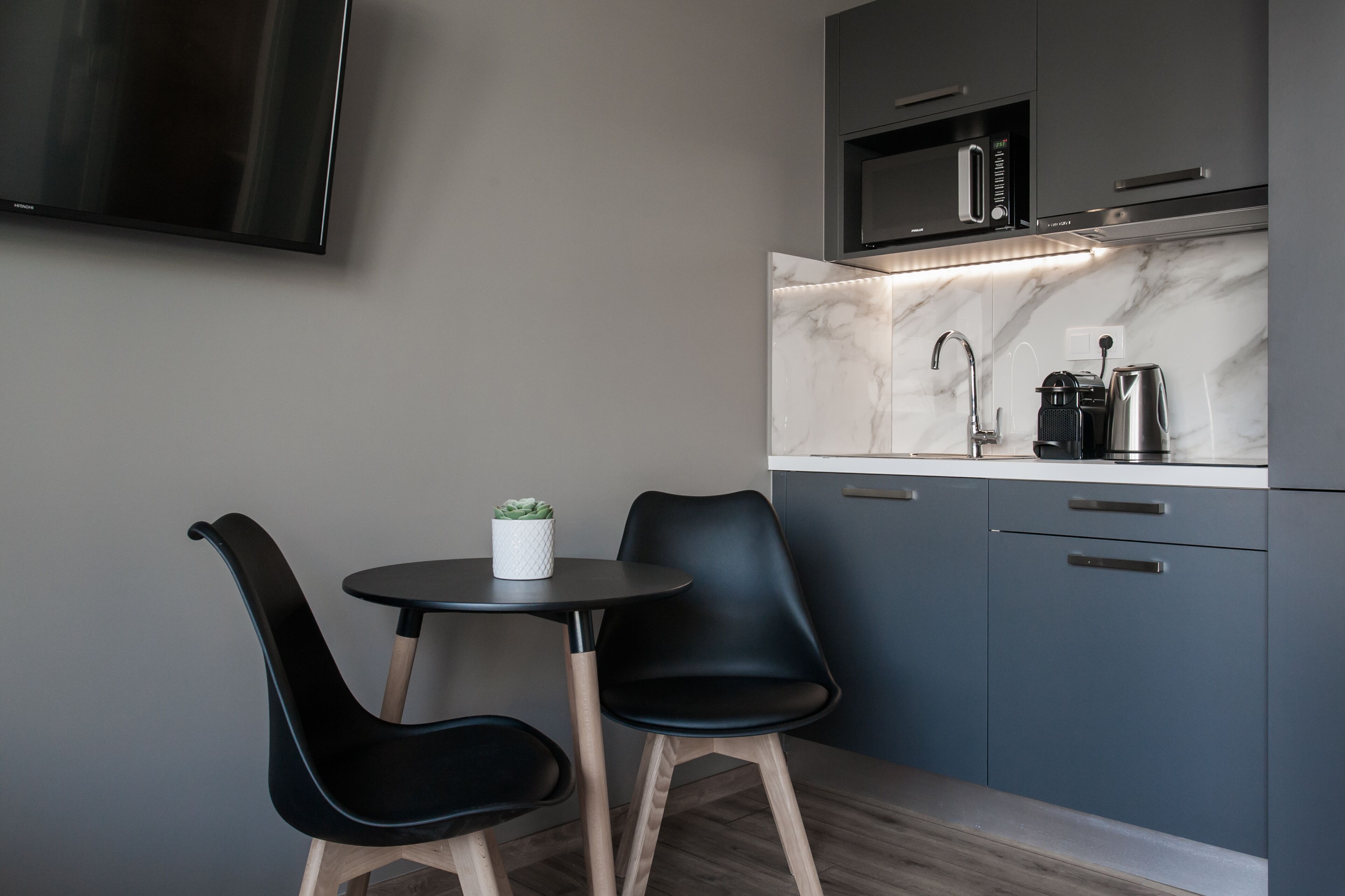 A modern kitchen with grey cabinets, a marble countertop, a black wall-mounted TV, and a small round table with two black chairs.