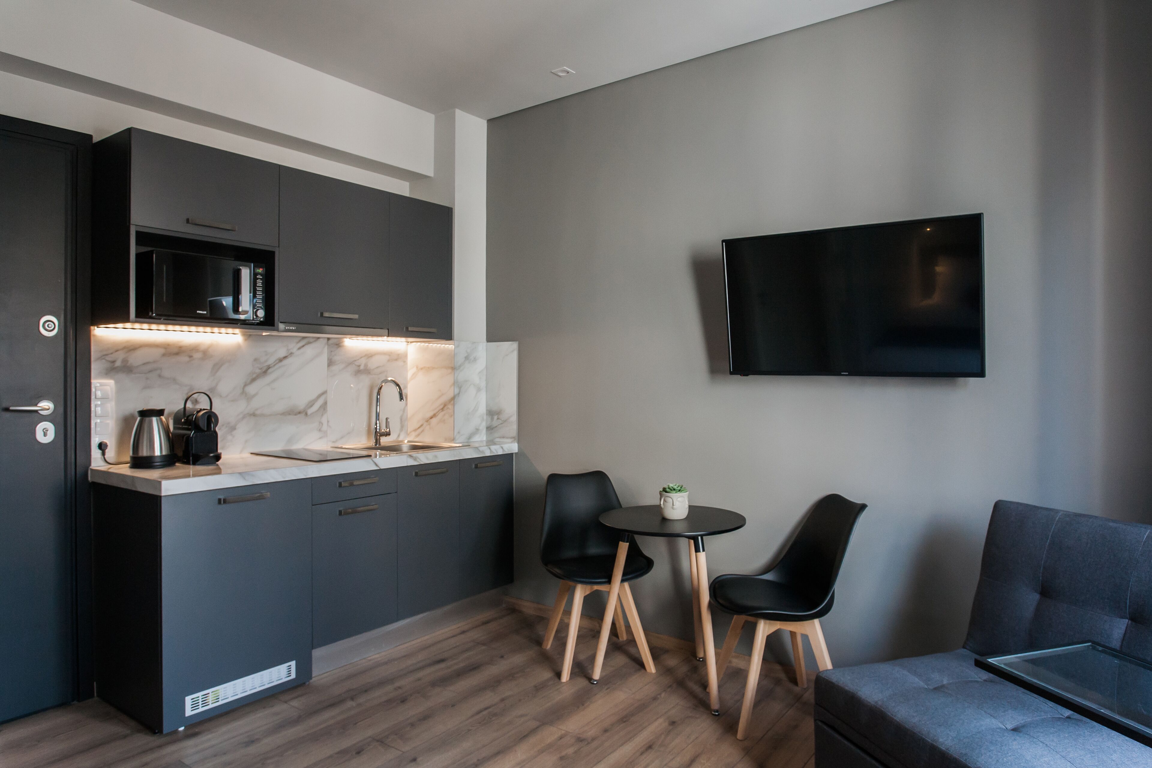 A modern kitchen with dark cabinetry, a marble countertop, and a built-in microwave. There is a small dining area with two chairs and a table, and a flat-screen TV mounted on the wall.