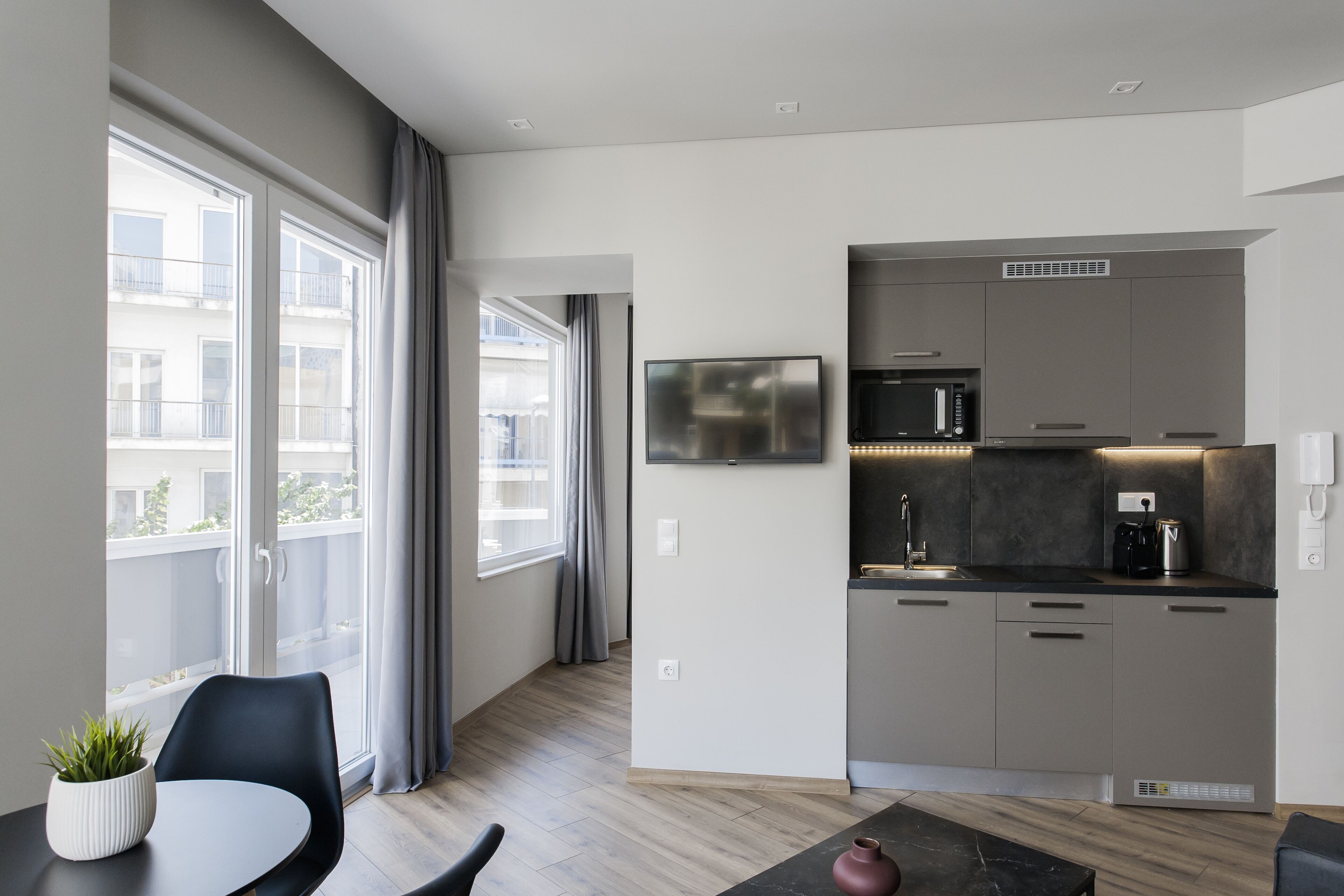 Een moderne keuken met grijze kastjes, een ingebouwde magnetron en een koffiezetapparaat. Er is een zithoek met een tafel en stoelen, en een potplant op de vensterbank.