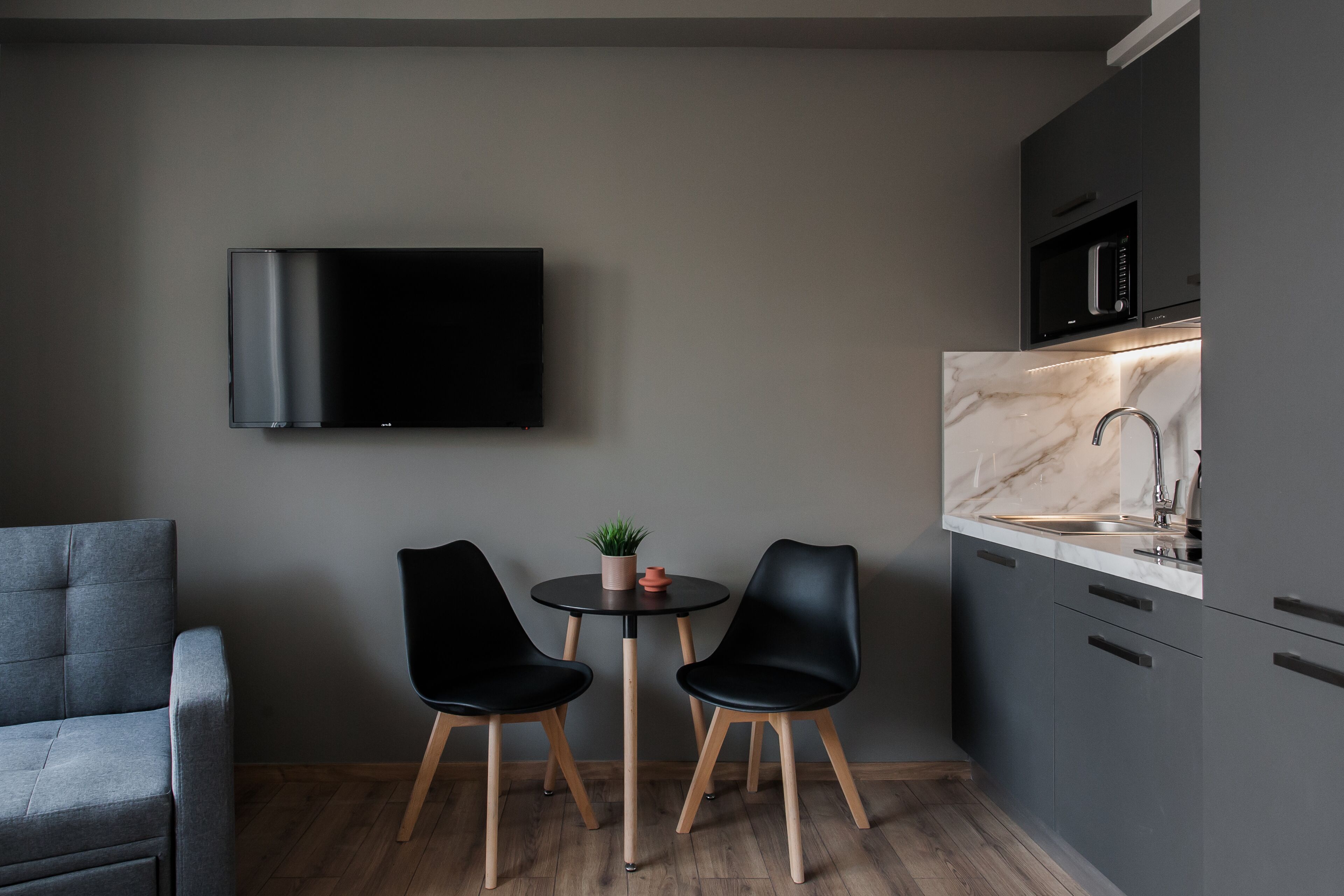 A modern kitchen with a flat-screen TV, two black chairs, a small table with a plant, and a marble countertop.