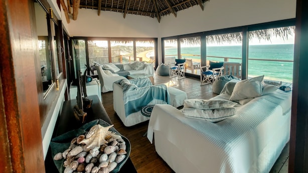 A room with a large window overlooking the sea, furnished with white sofas and chairs, a wooden deck, and a thatched roof. - The Beach House at Praia da Rocha (Inhambane)