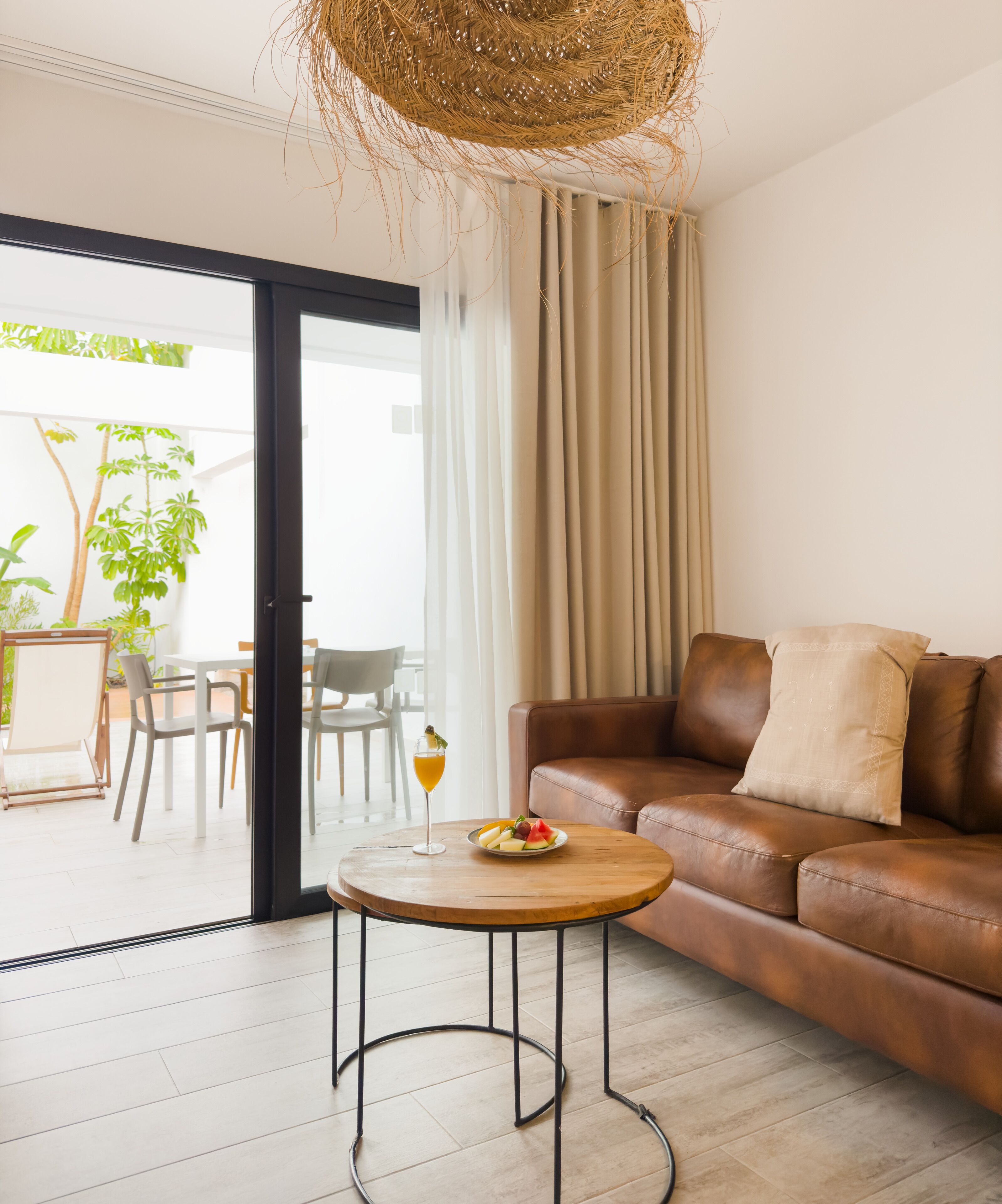 Ein modernes Wohnzimmer mit einem braunen Ledersofa, einem runden Couchtisch mit Obst und Getränken und einem großen Fenster mit Vorhängen.