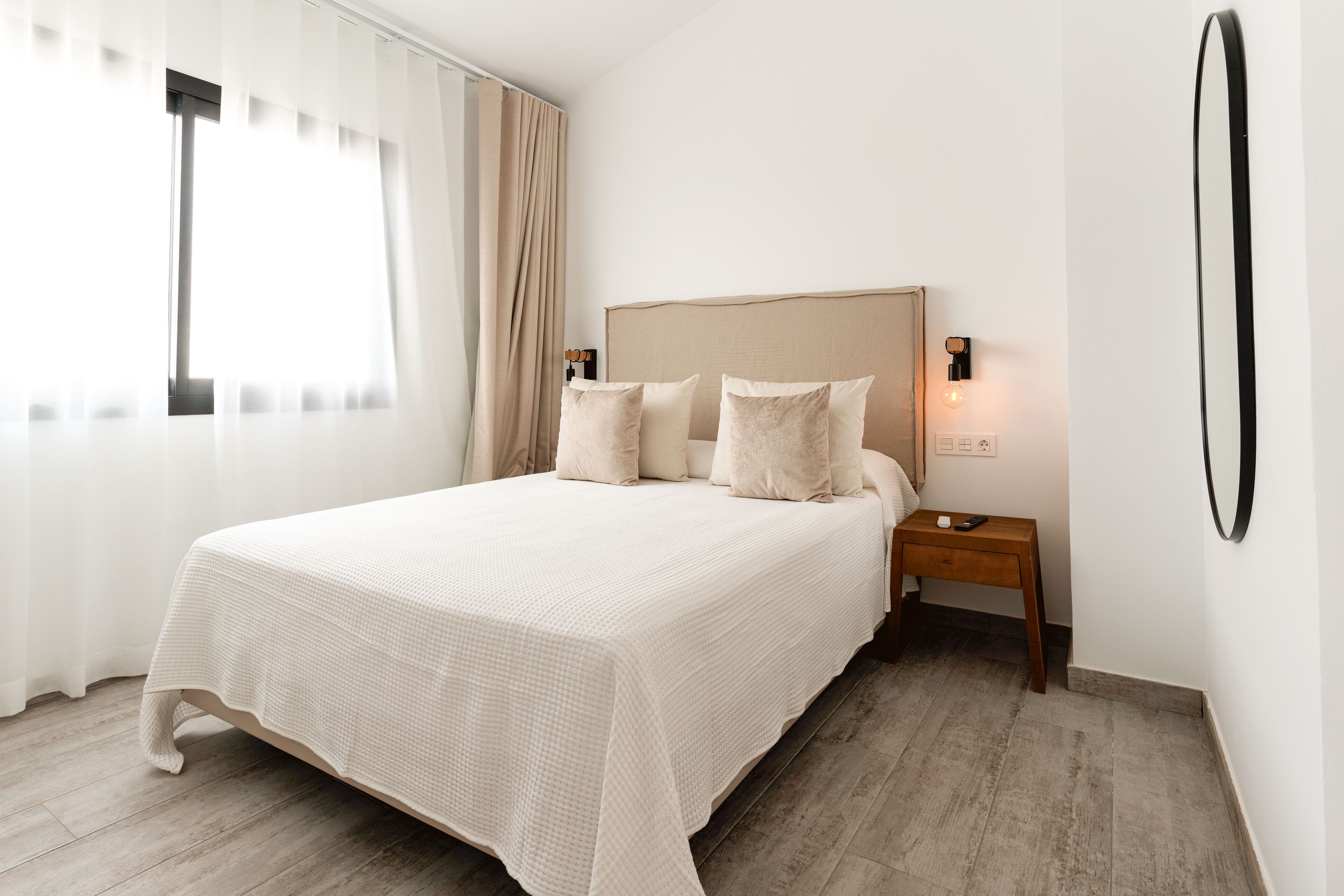Una cama bien hecha con una colcha color beige y almohadas, una mesita de noche de madera con una lámpara y un espejo grande en la pared.