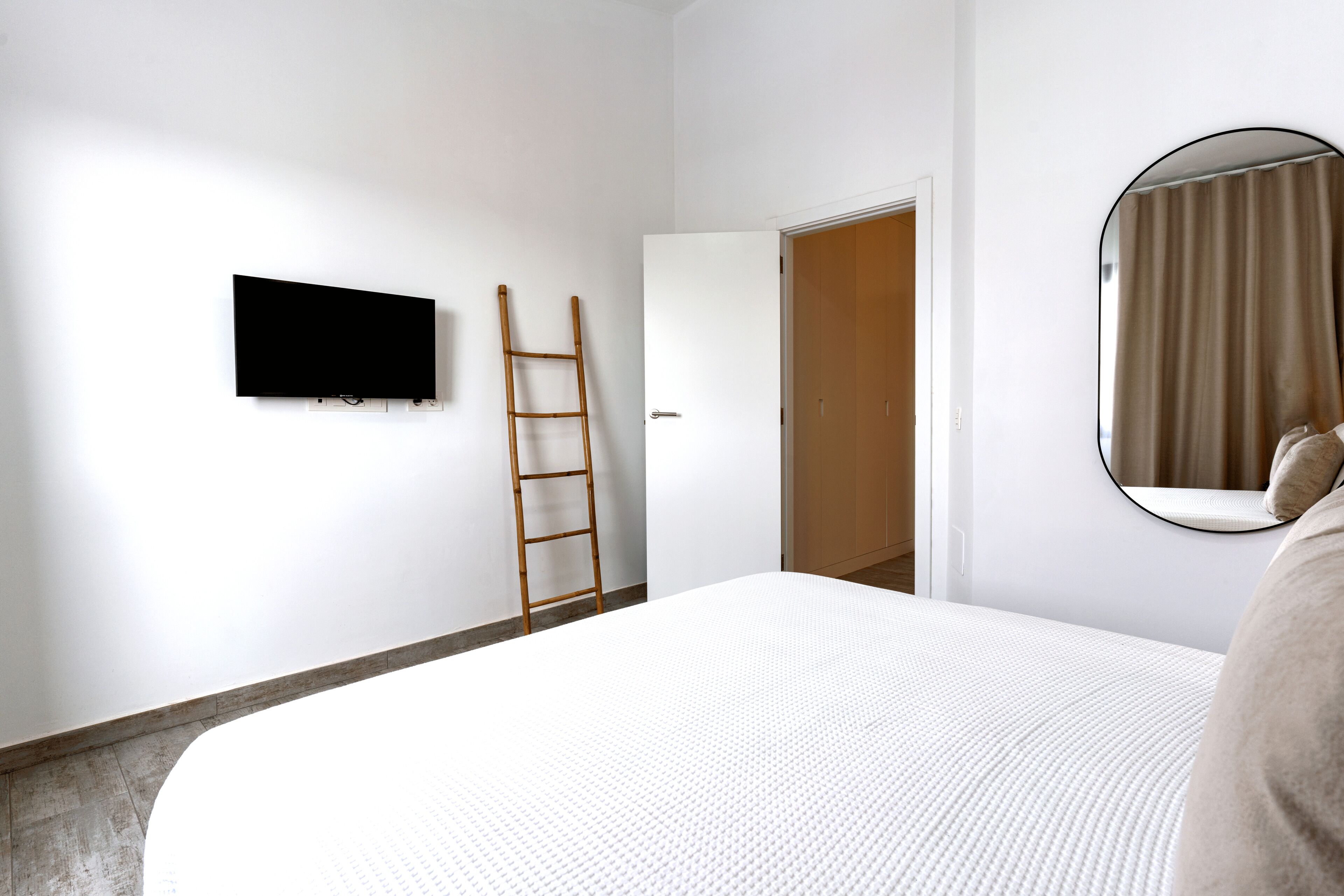 A bedroom with a bed, a TV mounted on the wall, a wooden ladder, a door, and a mirror with a curtain.