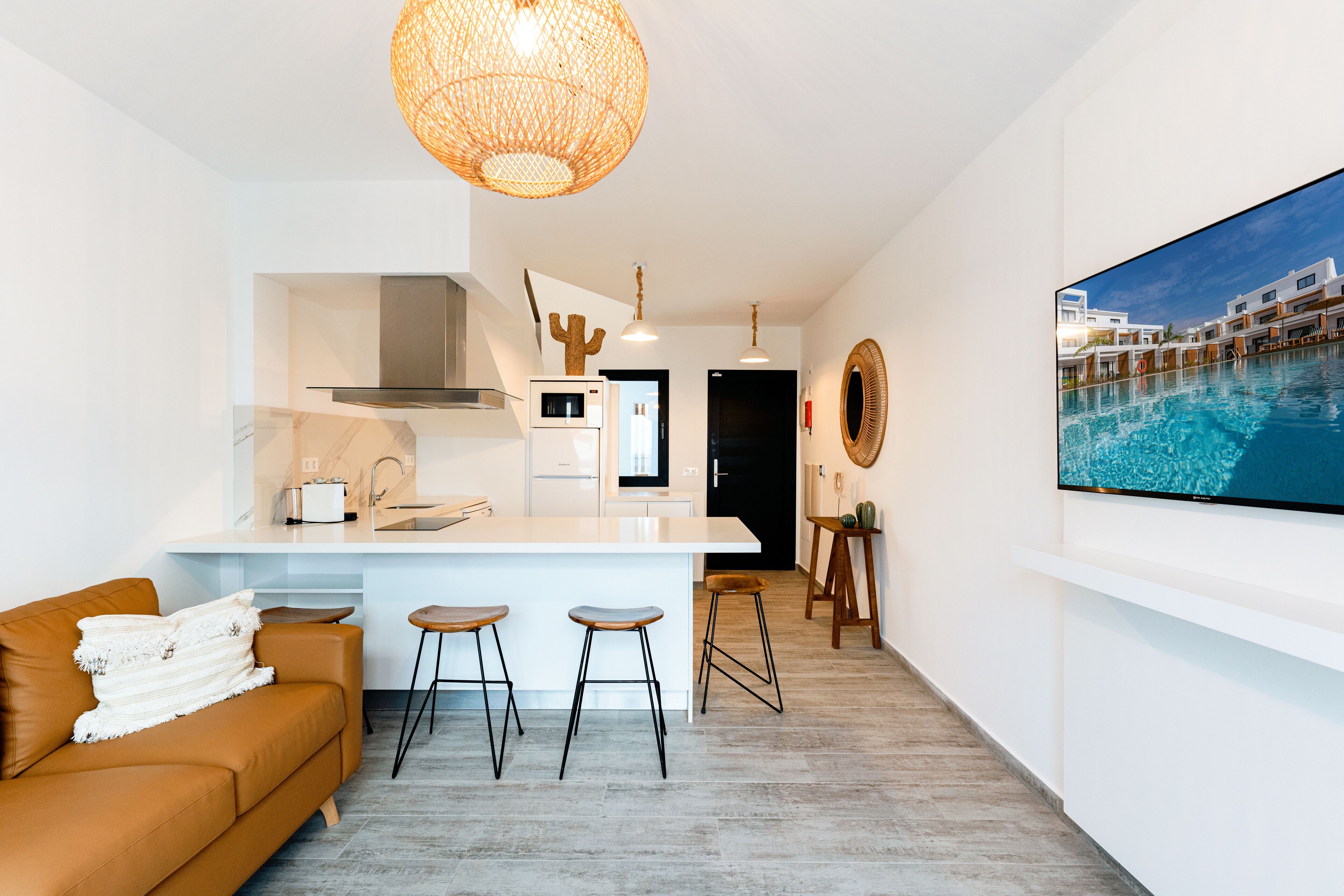 A modern kitchen with white cabinetry, a central island with stools, a hanging light fixture, and a large TV screen displaying a pool scene.