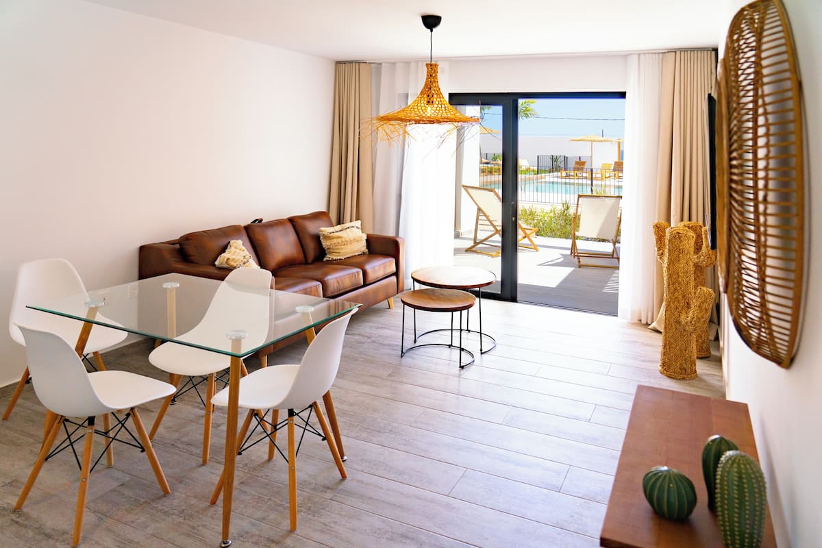 Een moderne woonkamer met een glazen eettafel, witte stoelen, een bruine leren bank en uitzicht op een buitenzwembad.