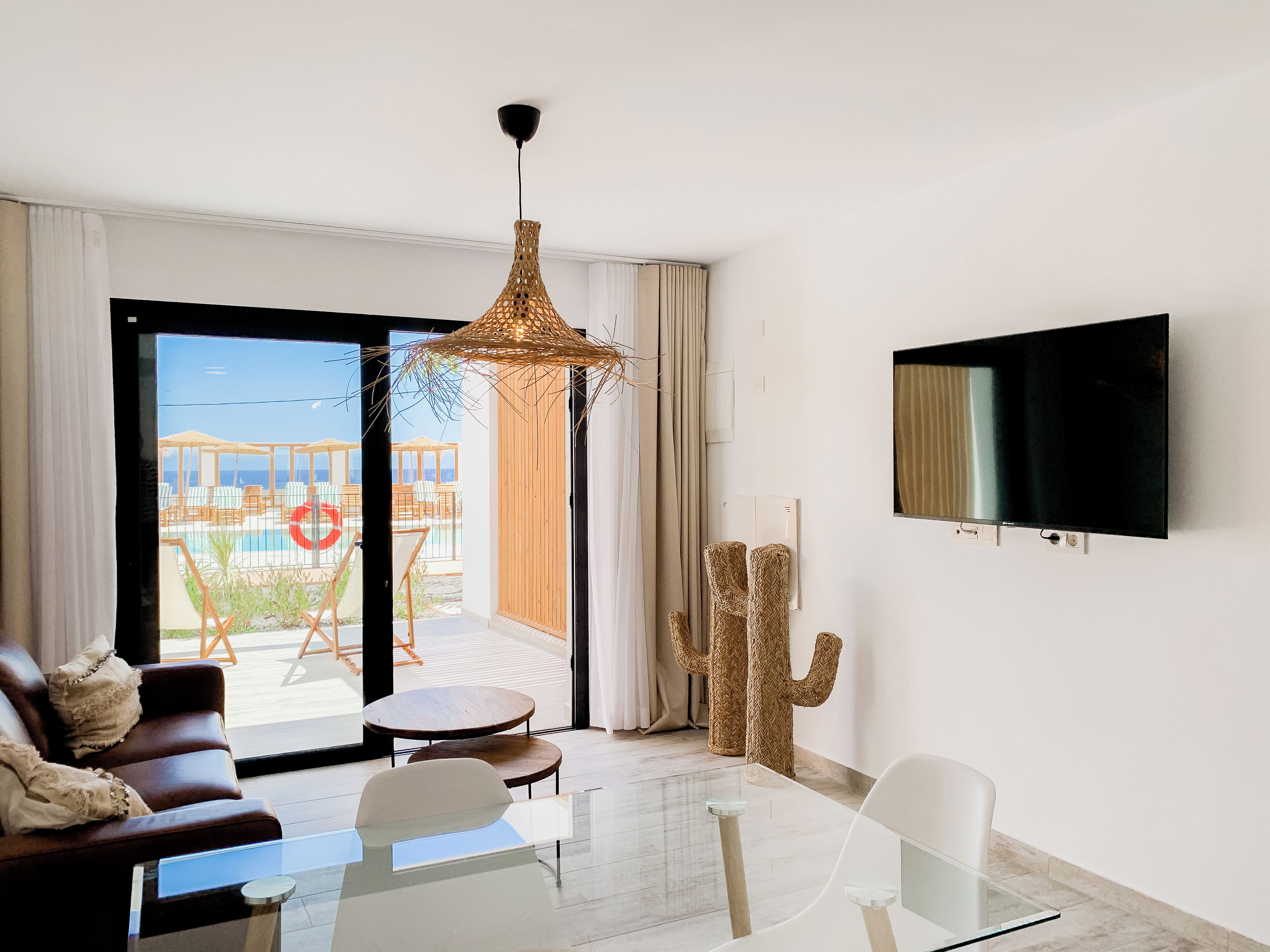 Un saló modern amb una taula de centre de vidre, un sofà de cuir marró, una butaca blanca, un gran televisor de pantalla plana fixat a la paret i vista a una zona de piscina exterior a través d'una porta corredissa de vidre.