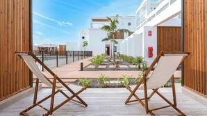 A poolside area with two wooden lounge chairs, a small palm tree, and a building with a white facade in the background. - Shambhala Fuerteventura (La Oliva)