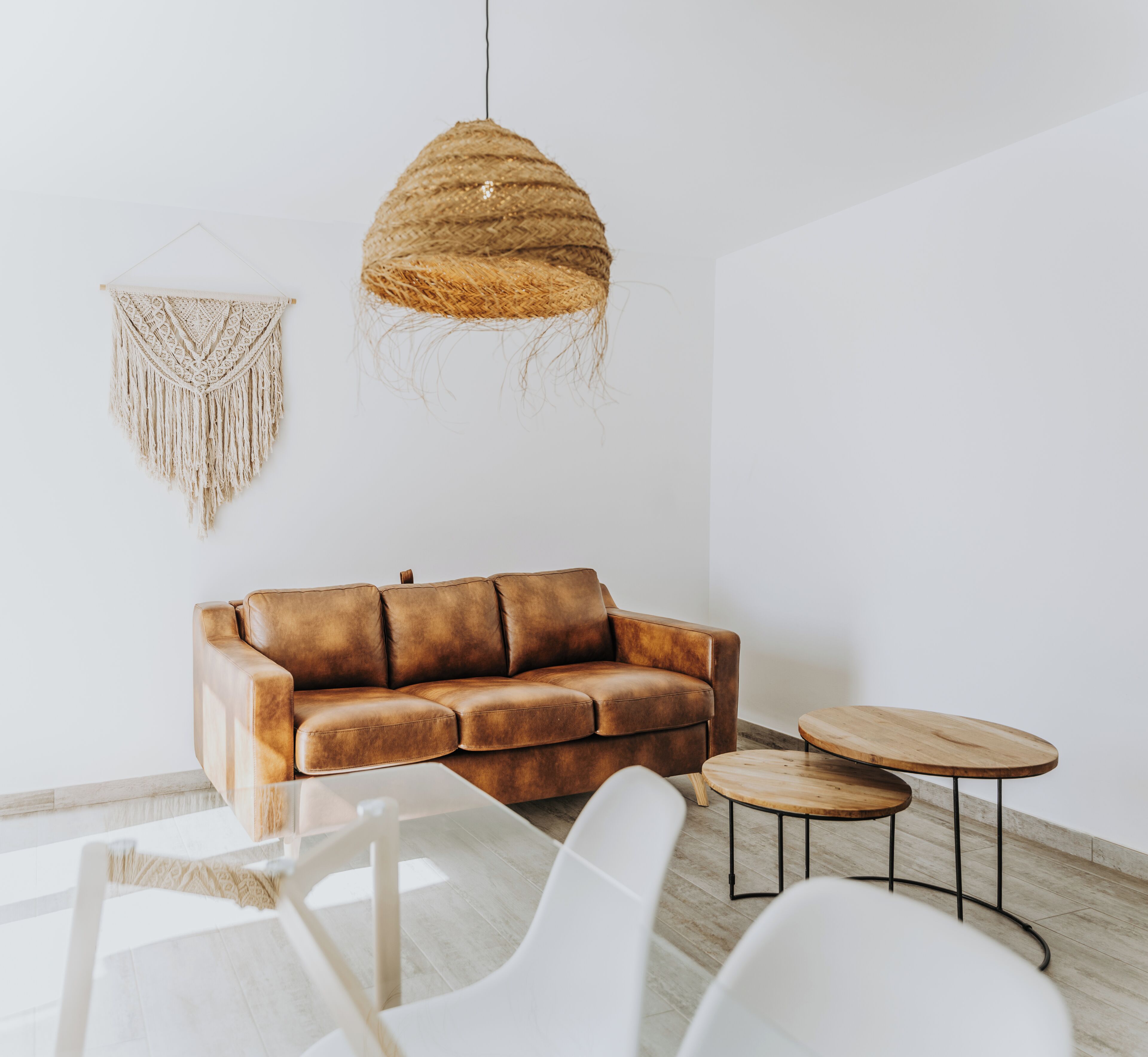 A modern living room with a leather sofa, two wooden side tables, a white chair, and a macramé wall hanging.