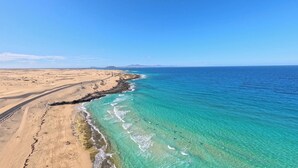 A coastal landscape with a clear blue sky, sandy beach, and turquoise waters. - Shambhala Fuerteventura (La Oliva)