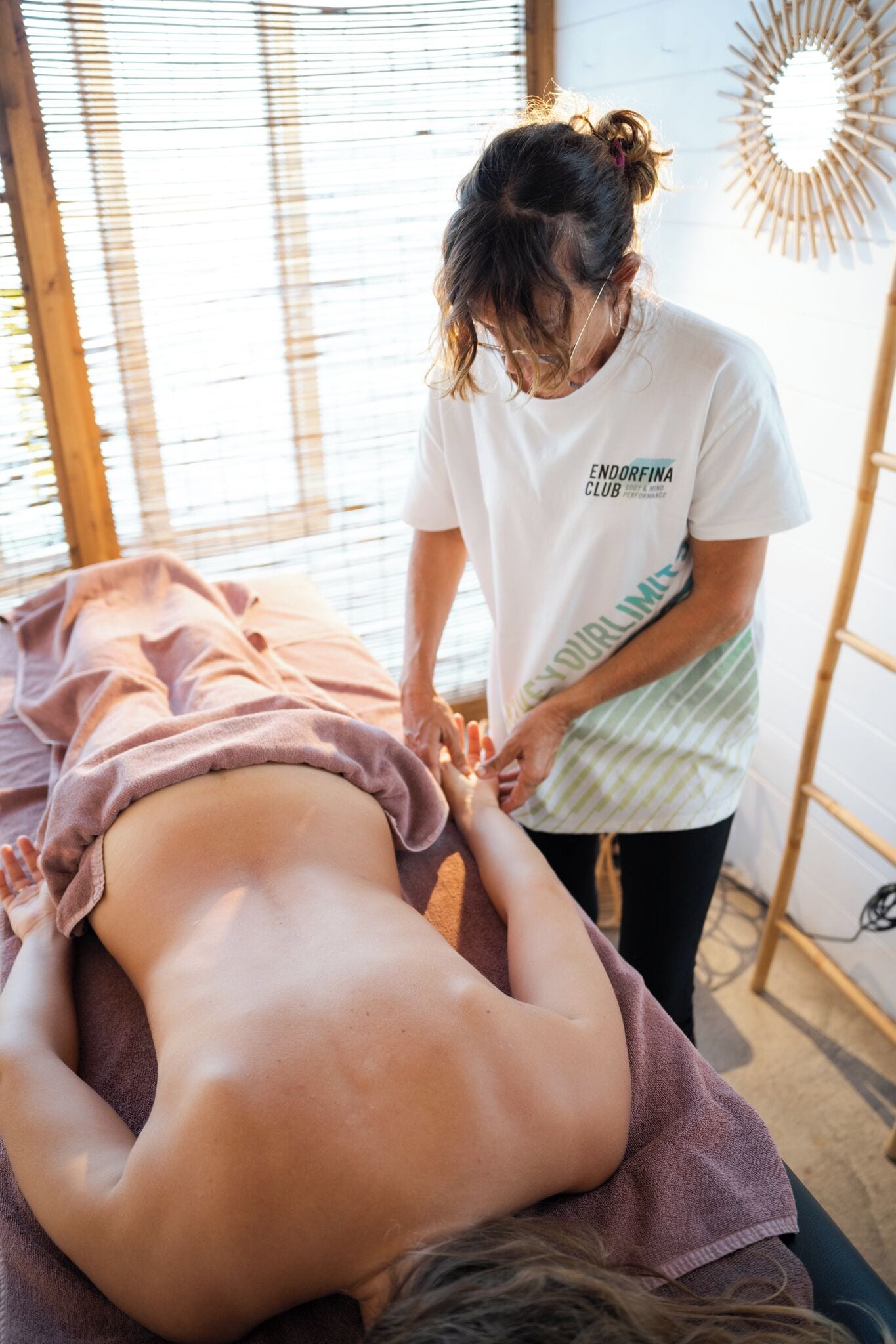 A massage therapy session in a room with a wooden floor, a large mirror, and a window with blinds.