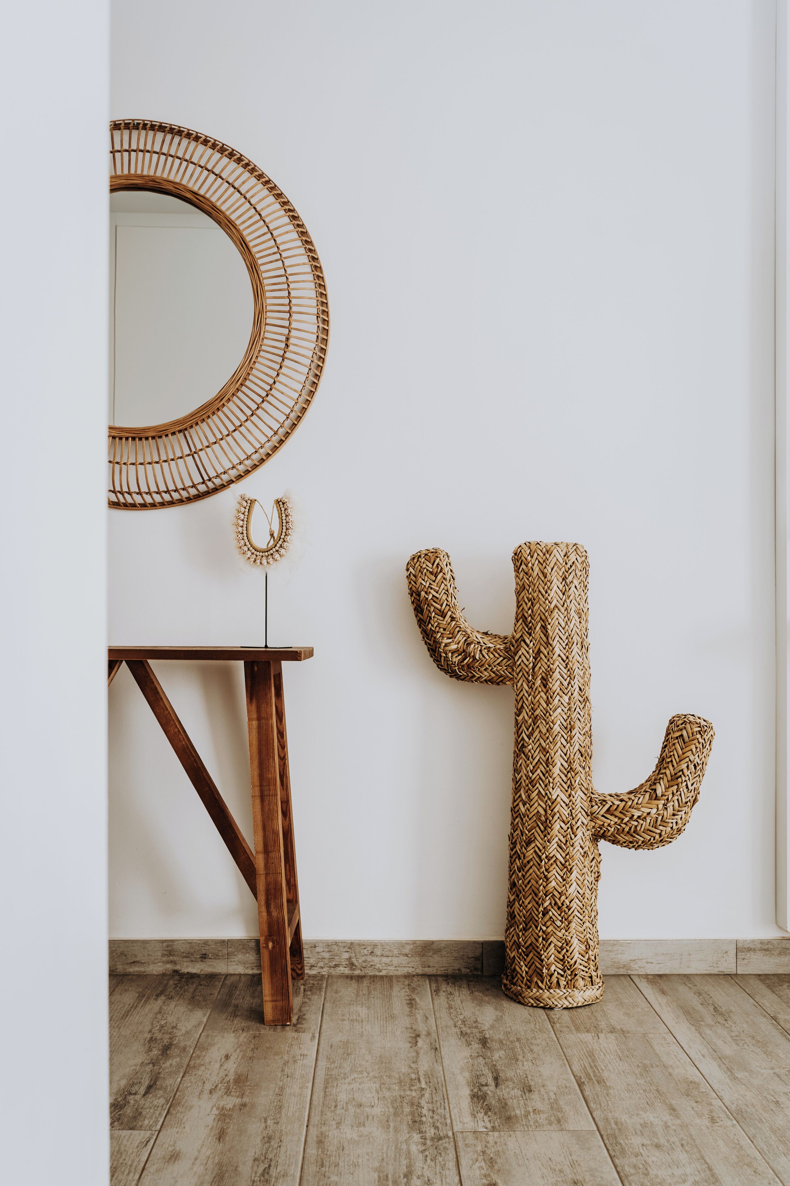 A minimalist room with a wooden floor, a round woven mirror, a small wooden table, and a woven cactus sculpture.