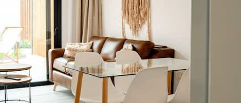 A modern living room with a glass dining table, white chairs, a brown leather sofa, and a macrame wall hanging.