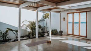 A spacious indoor area with a concrete floor, wooden ceiling beams, and large windows offering a view of the sea. - Shambhala Fuerteventura (La Oliva)