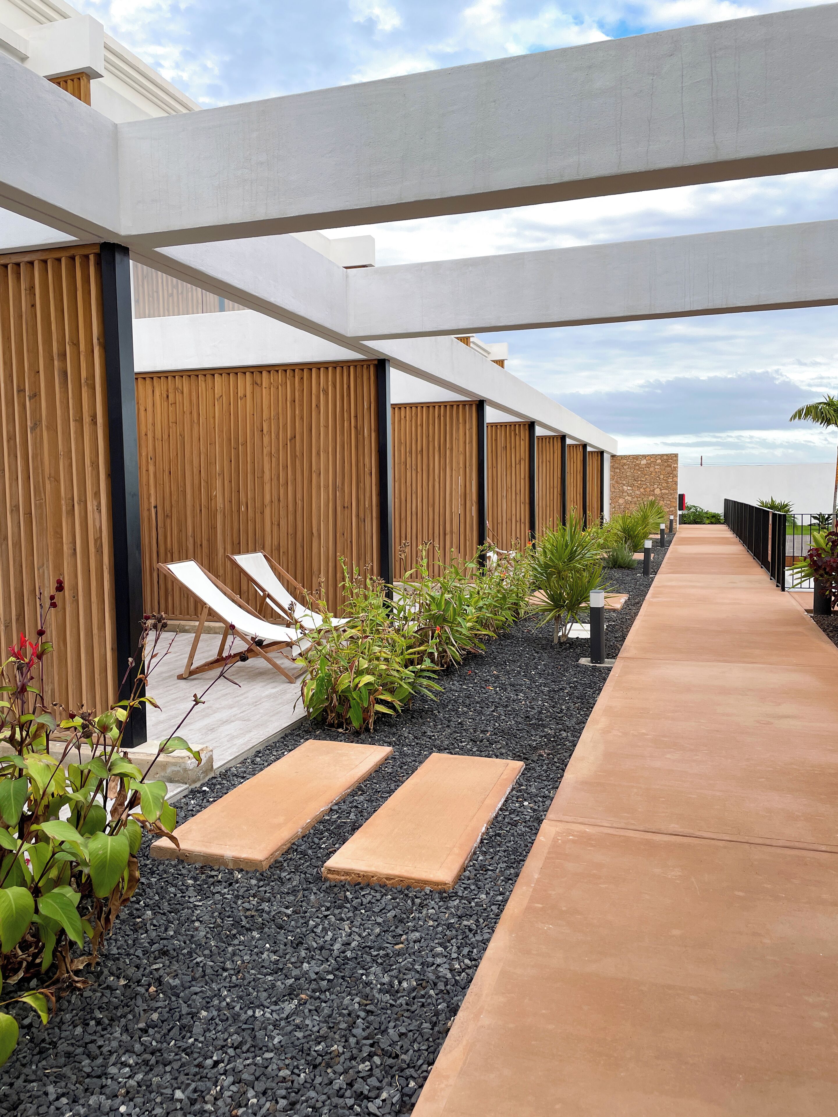 A modern outdoor lounge area with wooden slatted walls, a concrete pathway, and lounge chairs.